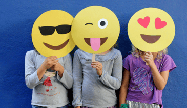 three young girls with paper emoji masks (from left to right: sunglasses emoji, tongue out emoji, and heart eyes emoji)