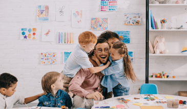 young male teacher with glasses being hugged by young students