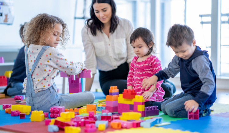 woman in white button down, sitting with young children on play mat as they play with large lego blocks